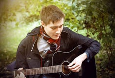 Un giovane tipo gioca una chitarra acustica nera, sedentesi nel parco immagine stock libera da diritti Un giovane tipo gioca una chitarra acustica nera, sedentesi nel parco immagine stock libera da diritti