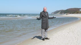 Un hombre joven elegante en una capa se coloca en la playa del mar, mira alrededor y sonríe - él es perdido él no sabe metrajes Un hombre joven elegante en una capa se coloca en la playa del mar, mira alrededor y sonríe - él es perdido él no sabe metrajes