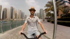 Un hombre joven en un sombrero, una camiseta rayada y pantalones cortos montando su bici en la 'promenade' entre los rascacielos almacen de metraje de vídeo Un hombre joven en un sombrero, una camiseta rayada y pantalones cortos montando su bici en la 'promenade' entre los rascacielos almacen de metraje de vídeo