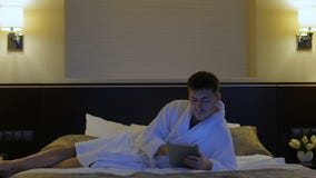 Un hombre joven está mintiendo en una cama en un hotel y mira en el teléfono, tableta almacen de metraje de vídeo Un hombre joven está mintiendo en una cama en un hotel y mira en el teléfono, tableta almacen de metraje de vídeo