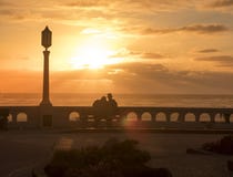 Un hombre joven y una mujer en el amor que se sienta en un banco en la costa y que disfruta de una puesta del sol hermosa fotos de archivo Un hombre joven y una mujer en el amor que se sienta en un banco en la costa y que disfruta de una puesta del sol hermosa fotos de archivo