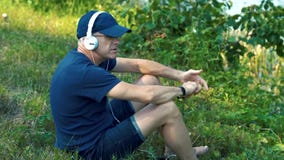 Un homme aux cheveux gris mince en écouteurs blancs, T-shirt bleu, chapeau et verres s'assied sur l'herbe verte sur la banque de clips vidéos Un homme aux cheveux gris mince en écouteurs blancs, T-shirt bleu, chapeau et verres s'assied sur l'herbe verte sur la banque de clips vidéos