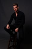 Un jeune, blanc type s'assied sur une chaise dans une chemise noire et un pantalon noir, contre un fond noir et regarder l'appare images stock Un jeune, blanc type s'assied sur une chaise dans une chemise noire et un pantalon noir, contre un fond noir et regarder l'appare images stock