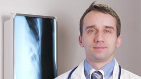 Un jeune docteur regarde l'appareil-photo et sourit Contre le rayon X accrochant de fond du patient Chemise avec un lien et banque de vidéos Un jeune docteur regarde l'appareil-photo et sourit Contre le rayon X accrochant de fond du patient Chemise avec un lien et banque de vidéos