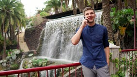 Un jeune homme d'affaires bel s'assied en parc et entretiens sur un smartphone - une rue et des cascades artificielles dans le tr banque de vidéos Un jeune homme d'affaires bel s'assied en parc et entretiens sur un smartphone - une rue et des cascades artificielles dans le tr banque de vidéos