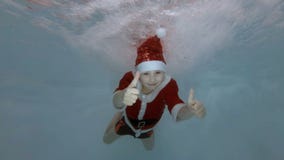 Un niño pequeño vestido como Santa Claus nada bajo el agua en los chorros de agua en la piscina con sus ojos abiertos, sonrisas, Un niño pequeño vestido como Santa Claus nada bajo el agua en los chorros de agua en la piscina con sus ojos abiertos, sonrisas,