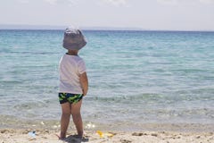 Un piccolo ragazzo timido con il cappello che sta nella sabbia vicino all'acqua e al lo immagine stock Un piccolo ragazzo timido con il cappello che sta nella sabbia vicino all'acqua e al lo immagine stock
