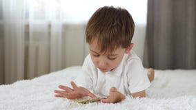 Un ragazzino sveglio esamina lo schermo dello smartphone mentre si trova sul letto I giochi da bambini sullo smartphone archivi video Un ragazzino sveglio esamina lo schermo dello smartphone mentre si trova sul letto I giochi da bambini sullo smartphone archivi video