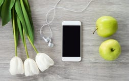 Un telefono bianco con le cuffie bianche, i tulipani bianchi e le bugie verdi delle mele su una tavola di legno bianca fotografia stock Un telefono bianco con le cuffie bianche, i tulipani bianchi e le bugie verdi delle mele su una tavola di legno bianca fotografia stock