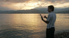 Un uomo in una camicia controlla i messaggi sulla compressa durante l'alba sulla spiaggia dell'oceano Colori meravigliosi del cie stock footage Un uomo in una camicia controlla i messaggi sulla compressa durante l'alba sulla spiaggia dell'oceano Colori meravigliosi del cie stock footage