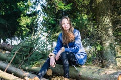 Una chica joven con el pelo rubio largo y una chaqueta del dril de algodón sienta en una clave un bosque conífero verde en un día foto de archivo Una chica joven con el pelo rubio largo y una chaqueta del dril de algodón sienta en una clave un bosque conífero verde en un día foto de archivo