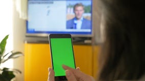 Una donna sta guardando la TV e sta tenendo uno smartphone con uno schermo verde Sulla manifestazione di TV le notizie Una donna sta guardando la TV e sta tenendo uno smartphone con uno schermo verde Sulla manifestazione di TV le notizie