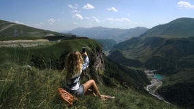 Una muchacha rubia joven se sienta en la hierba en un fondo de montañas con una mochila y toma las imágenes de las montañas almacen de metraje de vídeo Una muchacha rubia joven se sienta en la hierba en un fondo de montañas con una mochila y toma las imágenes de las montañas almacen de metraje de vídeo