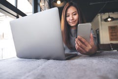 Una mujer asiática hermosa que sostiene y que mira el teléfono móvil mientras que usa el ordenador portátil en café imagen de archivo Una mujer asiática hermosa que sostiene y que mira el teléfono móvil mientras que usa el ordenador portátil en café imagen de archivo