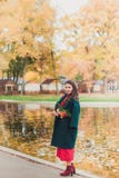 Una mujer joven camina en el parque del otoño Mujer morena que lleva una capa verde y un vestido rojo fotografía de archivo libre de regalías Una mujer joven camina en el parque del otoño Mujer morena que lleva una capa verde y un vestido rojo fotografía de archivo libre de regalías