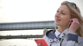 Una mujer rubia hermosa joven escucha la música en su smartphone usando los auriculares Una mujer joven disfruta de un paseo adel almacen de metraje de vídeo Una mujer rubia hermosa joven escucha la música en su smartphone usando los auriculares Una mujer joven disfruta de un paseo adel almacen de metraje de vídeo