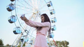 Una ragazza con capelli lunghi in un vestito lungo rosa fa il selfie facendo uso di uno smartphone mentre sta vicino alla ruota p stock footage Una ragazza con capelli lunghi in un vestito lungo rosa fa il selfie facendo uso di uno smartphone mentre sta vicino alla ruota p stock footage