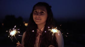 Una ragazza con capelli scuri lunghi sta contro lo sfondo della città alla notte e tiene i fuochi d'artificio in sue mani archivi video Una ragazza con capelli scuri lunghi sta contro lo sfondo della città alla notte e tiene i fuochi d'artificio in sue mani archivi video