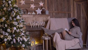 Una ragazza con capelli scuri lunghi sta leggendo un libro mentre si sedeva in una poltrona vicino ad un albero di Natale 4K stock footage Una ragazza con capelli scuri lunghi sta leggendo un libro mentre si sedeva in una poltrona vicino ad un albero di Natale 4K stock footage