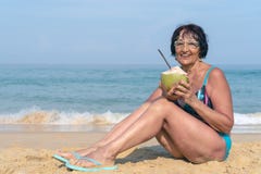Une femme agée avec les cheveux noirs s'assied par la mer un jour ensoleillé Une femme dans un maillot de bain avec une noix de c images libres de droits Une femme agée avec les cheveux noirs s'assied par la mer un jour ensoleillé Une femme dans un maillot de bain avec une noix de c images libres de droits