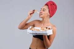 Une fille est dans les sous-vêtements noirs et est enveloppée dans une serviette sur le chef, mangeant un morceau de pizza appéti photographie stock libre de droits Une fille est dans les sous-vêtements noirs et est enveloppée dans une serviette sur le chef, mangeant un morceau de pizza appéti photographie stock libre de droits