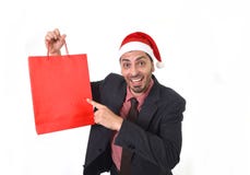 Ung attraktiv affärsman i juljultomtenhatten som rymmer och pekar den röda shoppingpåsen i den December och för nytt år försäljni arkivbild Ung attraktiv affärsman i juljultomtenhatten som rymmer och pekar den röda shoppingpåsen i den December och för nytt år försäljni arkivbild