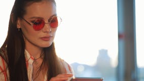 Ung attraktiv kvinna i röd solglasögon genom att använda minnestavladatoren med pekskärmen i ett kafé Härlig flicka i flygplats e stock video Ung attraktiv kvinna i röd solglasögon genom att använda minnestavladatoren med pekskärmen i ett kafé Härlig flicka i flygplats e stock video