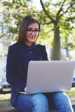 Ung studentflicka i exponeringsglas som sitter på universitetsområdebänken medan bruksbärbar datorgrej under hennes gruppavbrott fotografering för bildbyråer Ung studentflicka i exponeringsglas som sitter på universitetsområdebänken medan bruksbärbar datorgrej under hennes gruppavbrott fotografering för bildbyråer
