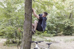 Unga roliga caucasian manklättringar upp till trädet med mässan eller fasa, den vita cykeln står ner under Vita trådlösa hörlurar royaltyfri fotografi Unga roliga caucasian manklättringar upp till trädet med mässan eller fasa, den vita cykeln står ner under Vita trådlösa hörlurar royaltyfri fotografi