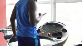 Uomo afroamericano che cammina sulla pedana mobile e che controlla il suo smartphone nella palestra archivi video Uomo afroamericano che cammina sulla pedana mobile e che controlla il suo smartphone nella palestra archivi video