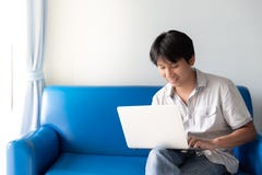 Uomo asiatico bello facendo uso del computer portatile per gli impianti e le attività online mentre sedendosi sul sofà blu immagini stock libere da diritti Uomo asiatico bello facendo uso del computer portatile per gli impianti e le attività online mentre sedendosi sul sofà blu immagini stock libere da diritti