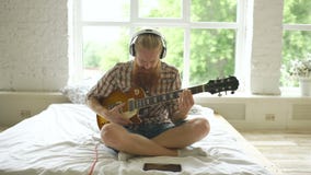 Uomo barbuto attraente in cuffie che si siedono sul letto che impara giocare chitarra facendo uso del computer della compressa in archivi video Uomo barbuto attraente in cuffie che si siedono sul letto che impara giocare chitarra facendo uso del computer della compressa in archivi video