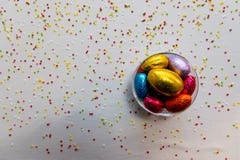 Uova di Pasqua variopinte del cioccolato in una ciotola trasparente con fondo bianco e coriandoli vaghi fotografia stock Uova di Pasqua variopinte del cioccolato in una ciotola trasparente con fondo bianco e coriandoli vaghi fotografia stock