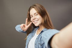 Uśmiechnięta brązowowłosa dziewczyna ubierająca w białej cajg koszula i koszulce pokazuje selfie i robi na szarości v śpiewa obraz stock Uśmiechnięta brązowowłosa dziewczyna ubierająca w białej cajg koszula i koszulce pokazuje selfie i robi na szarości v śpiewa obraz stock