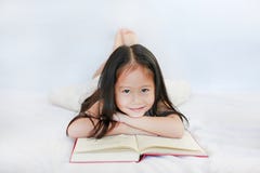 Uśmiechnięta mała Azjatycka dziecko dziewczyna z hardcover książki lying on the beach na łóżkowej i przyglądającej kamerze nad bi obrazy stock Uśmiechnięta mała Azjatycka dziecko dziewczyna z hardcover książki lying on the beach na łóżkowej i przyglądającej kamerze nad bi obrazy stock
