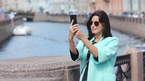 Uśmiechnięta modnej kobiety turystyczna bierze fotografia używać smartphone pozycję na moście nad rzeką zdjęcie wideo Uśmiechnięta modnej kobiety turystyczna bierze fotografia używać smartphone pozycję na moście nad rzeką zdjęcie wideo