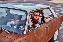 Varón barbudo en las gafas de sol vestidas en la chaqueta de cuero marrón que conduce un coche retro fotografía de archivo libre de regalías Varón barbudo en las gafas de sol vestidas en la chaqueta de cuero marrón que conduce un coche retro fotografía de archivo libre de regalías