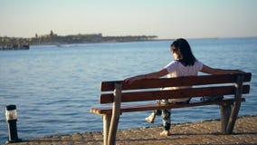Verano, puesta del sol, terraplén por el mar, una mujer morena joven se sienta en un banco, parte posterior, admirando una puesta almacen de video Verano, puesta del sol, terraplén por el mar, una mujer morena joven se sienta en un banco, parte posterior, admirando una puesta almacen de video
