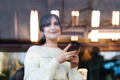 Vista através do caso de vidro Retrato da jovem mulher de sorriso com o smartphone em suas m?os, sentando-se no caf? da tabela, o fotografia de stock royalty free Vista através do caso de vidro Retrato da jovem mulher de sorriso com o smartphone em suas m?os, sentando-se no caf? da tabela, o fotografia de stock royalty free