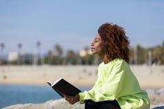 Vista laterale di giovane donna riccia di afro che si siede su un frangiflutti che tiene un libro mentre sorridendo e distogliend fotografia stock libera da diritti Vista laterale di giovane donna riccia di afro che si siede su un frangiflutti che tiene un libro mentre sorridendo e distogliend fotografia stock libera da diritti