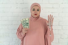 Vit kvinna med blåa ögon i en rosa hijab som rymmer en radband och dollar på en vit bakgrund arkivbild Vit kvinna med blåa ögon i en rosa hijab som rymmer en radband och dollar på en vit bakgrund arkivbild