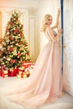 Vrouw in een lange roomkleurige kleding, die zich dichtbij de Kerstboom en de deur bevinden Het luxueuze blonde in avondjurk vier stock foto's Vrouw in een lange roomkleurige kleding, die zich dichtbij de Kerstboom en de deur bevinden Het luxueuze blonde in avondjurk vier stock foto's