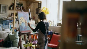 Vue arrière de jeune fille de peintre dans le tablier peignant toujours le tableau de la vie sur la toile dans classe de l'art banque de vidéos Vue arrière de jeune fille de peintre dans le tablier peignant toujours le tableau de la vie sur la toile dans classe de l'art banque de vidéos