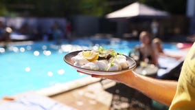 Waiter carries oysters and scallops on tray, with slices of lemon with a swimming pool on background at luxury hotel or
