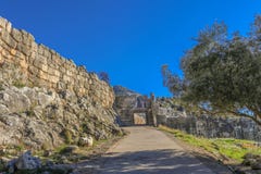 Weergeven van Leeuwenpoort in oude Mycenae Griekenland van onderaan de heuvel die die de stoep tonen - door een olijfboom in de s stock afbeeldingen Weergeven van Leeuwenpoort in oude Mycenae Griekenland van onderaan de heuvel die die de stoep tonen - door een olijfboom in de s stock afbeeldingen