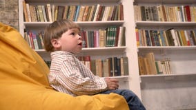 Weinig leuke jong geitjezitting op stoel en het vangen van bal van somebody, boekenrekkenachtergrond stock footage Weinig leuke jong geitjezitting op stoel en het vangen van bal van somebody, boekenrekkenachtergrond stock footage