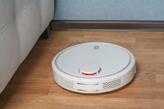Weißer RoboterStaubsauger läuft in nahes Ecksofa auf hölzernem Parkettboden Moderne intelligente Reinigungstechnologiehaushaltung stockfotos Weißer RoboterStaubsauger läuft in nahes Ecksofa auf hölzernem Parkettboden Moderne intelligente Reinigungstechnologiehaushaltung stockfotos