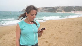 Woman on beach listening to music on headphones from smart phone. Nazare, Portugal. Slow motion stock footage Woman on beach listening to music on headphones from smart phone. Nazare, Portugal. Slow motion stock footage