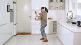 Woman dancing while listening music in kitchen. Beautiful young woman holding mobile phone to listen music with headphones while singing and dancing in the Woman dancing while listening music in kitchen. Beautiful young woman holding mobile phone to listen music with headphones while singing and dancing in the