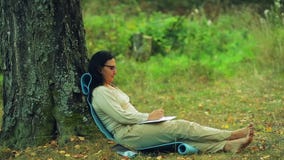A woman in glasses barefoot sits under a tree in the park and draws a pencil in a notebook